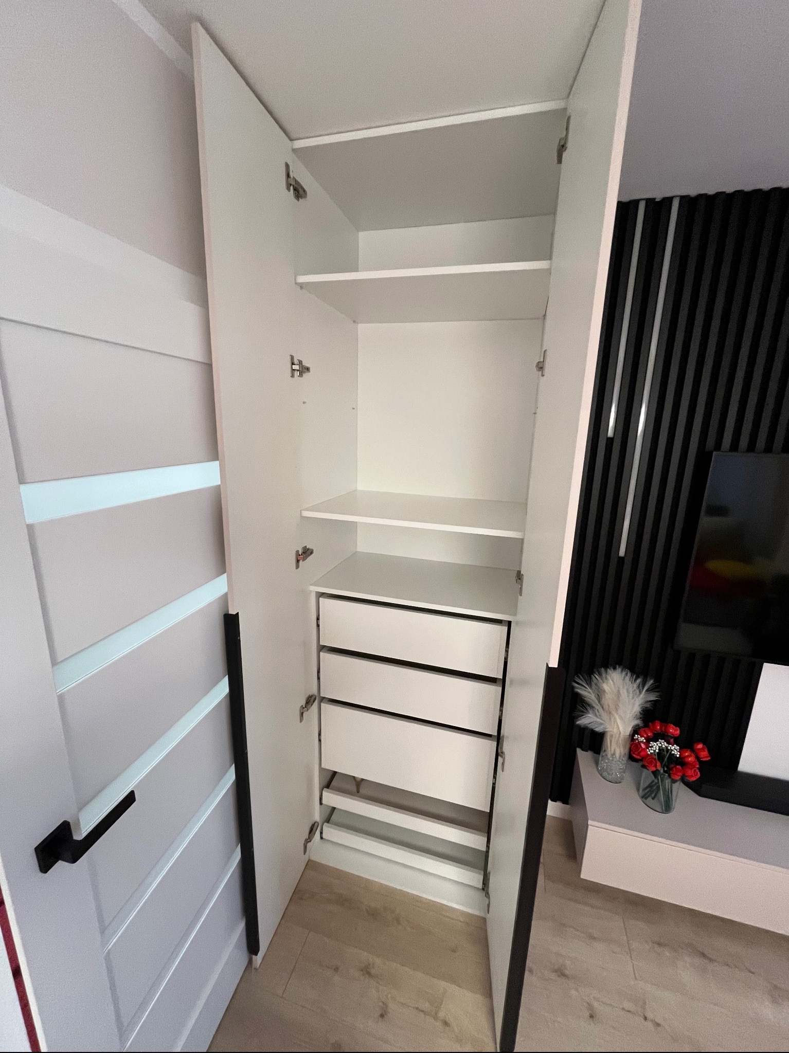 Wnętrze białej szafy wnękowej z otwartymi drzwiami, widoczne półki i szuflady, nowoczesny design, czarne akcenty na drzwiach i ścianie w tle.