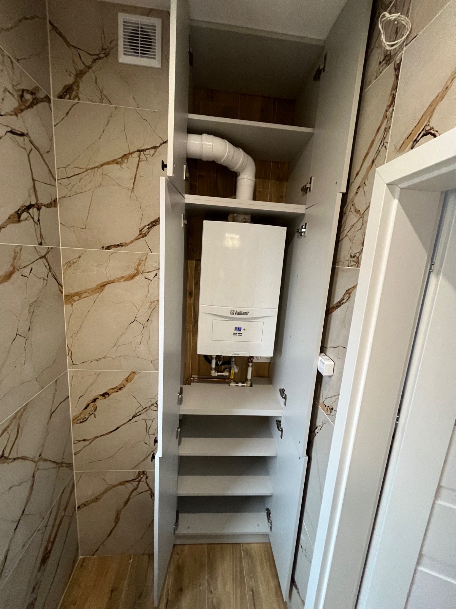 Biała szafa wnękowa z otwartymi drzwiami, ukrywająca piec gazowy Vaillant, widoczne rury, półki i marmurowe płytki na ścianie.