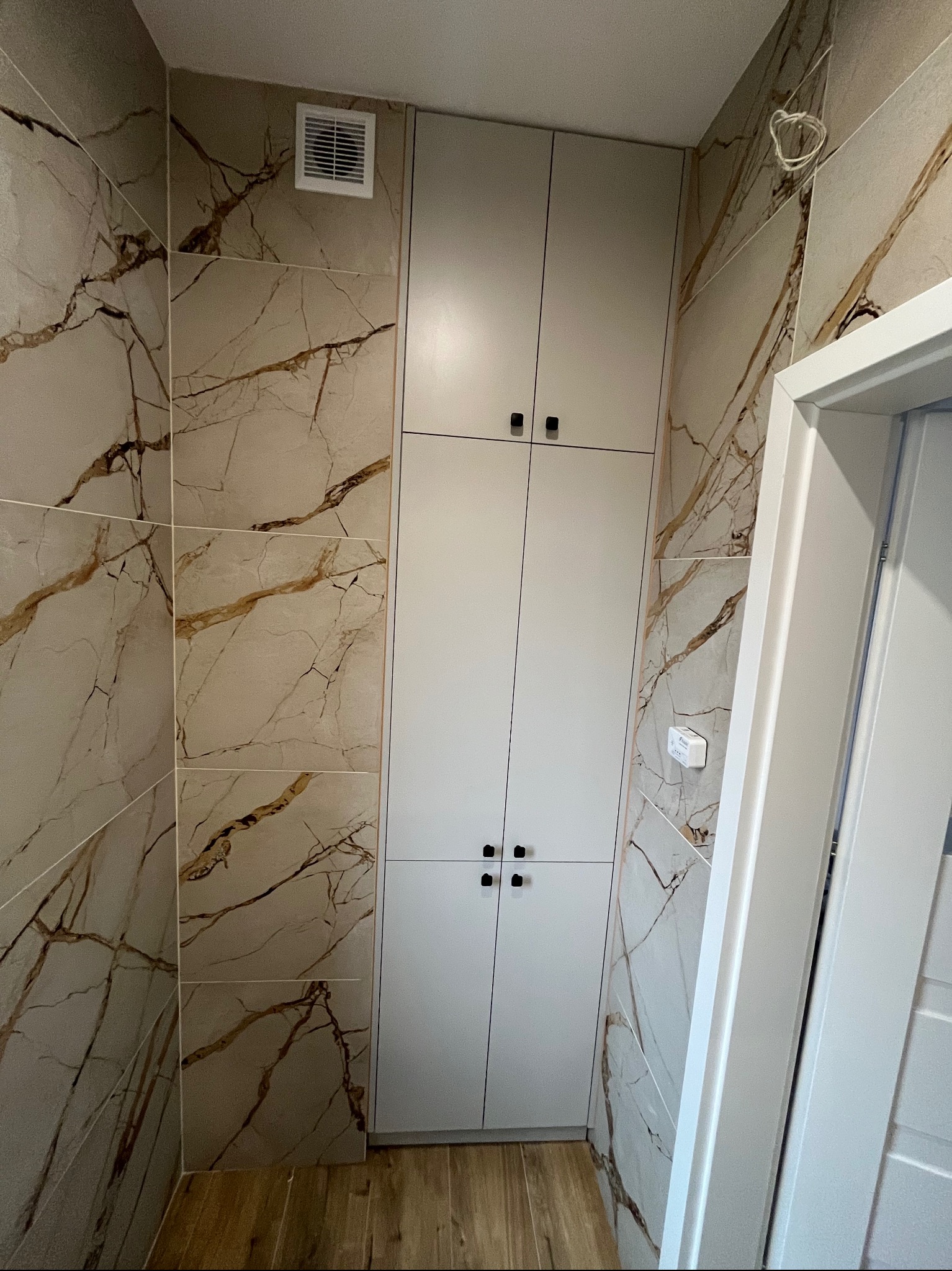 Wysoka, zabudowana szafa wnękowa z czterema drzwiami i czarnymi uchwytami, w łazience wykończonej płytkami imitującymi marmur z brązowymi żyłami, widoczna kratka wentylacyjna na górze.