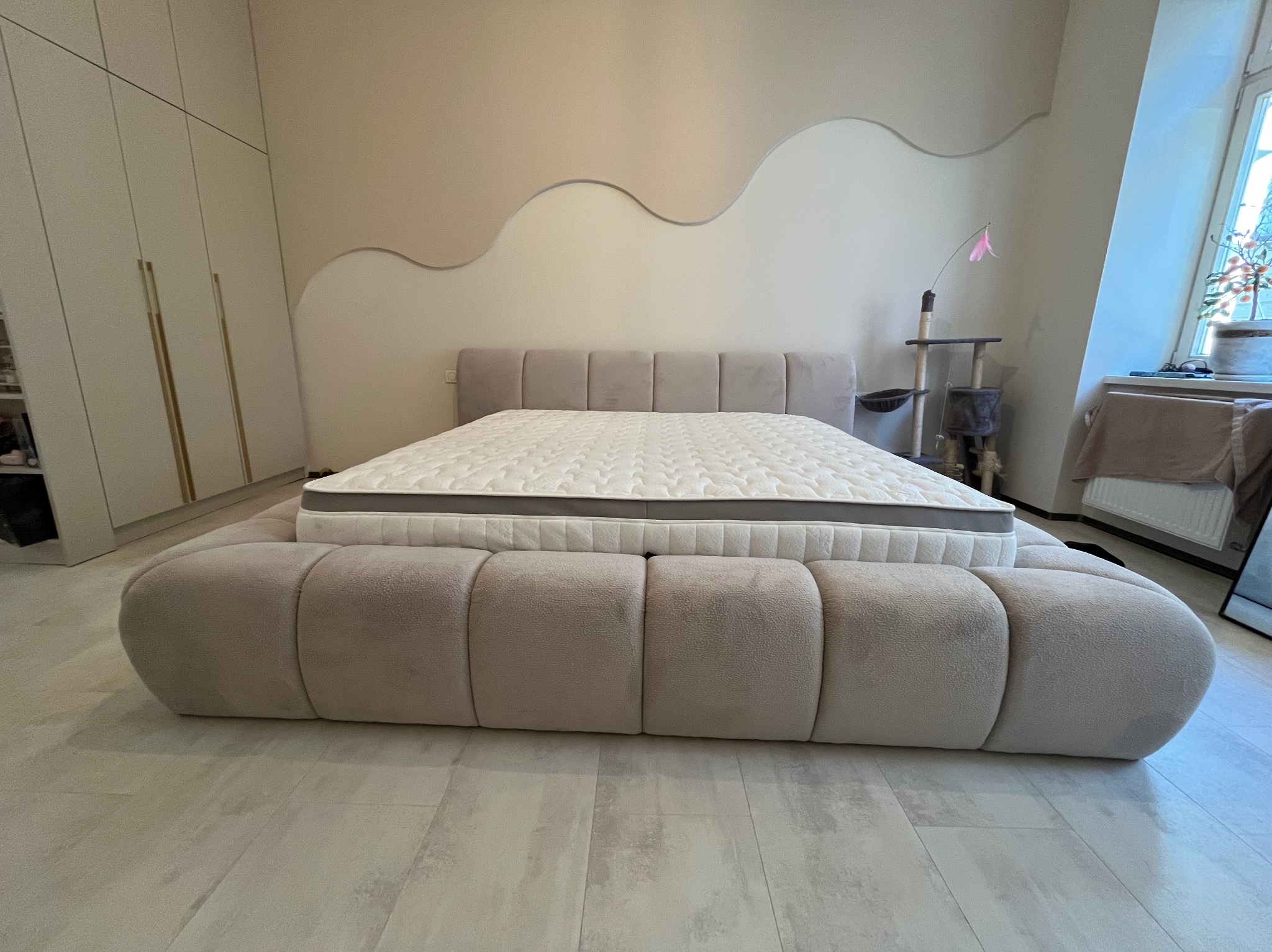 Beżowe łóżko tapicerowane z niskim wezgłowiem w sypialni z dekoracyjną falą na ścianie i jasnymi szafami w tle.