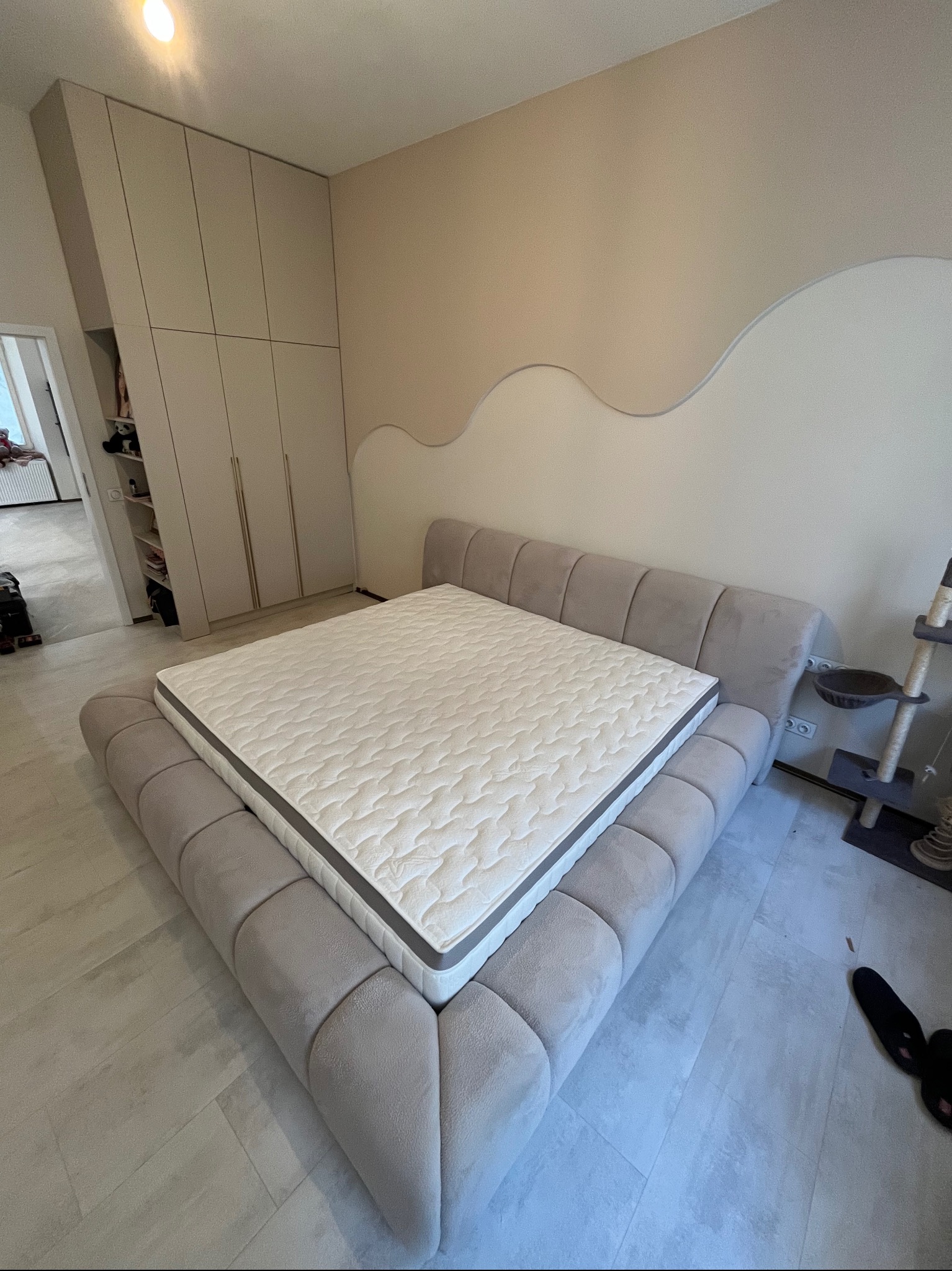 Beżowe łóżko tapicerowane z materacem w sypialni z dużą szafą wnękową i dekoracyjną ścianą w tle.