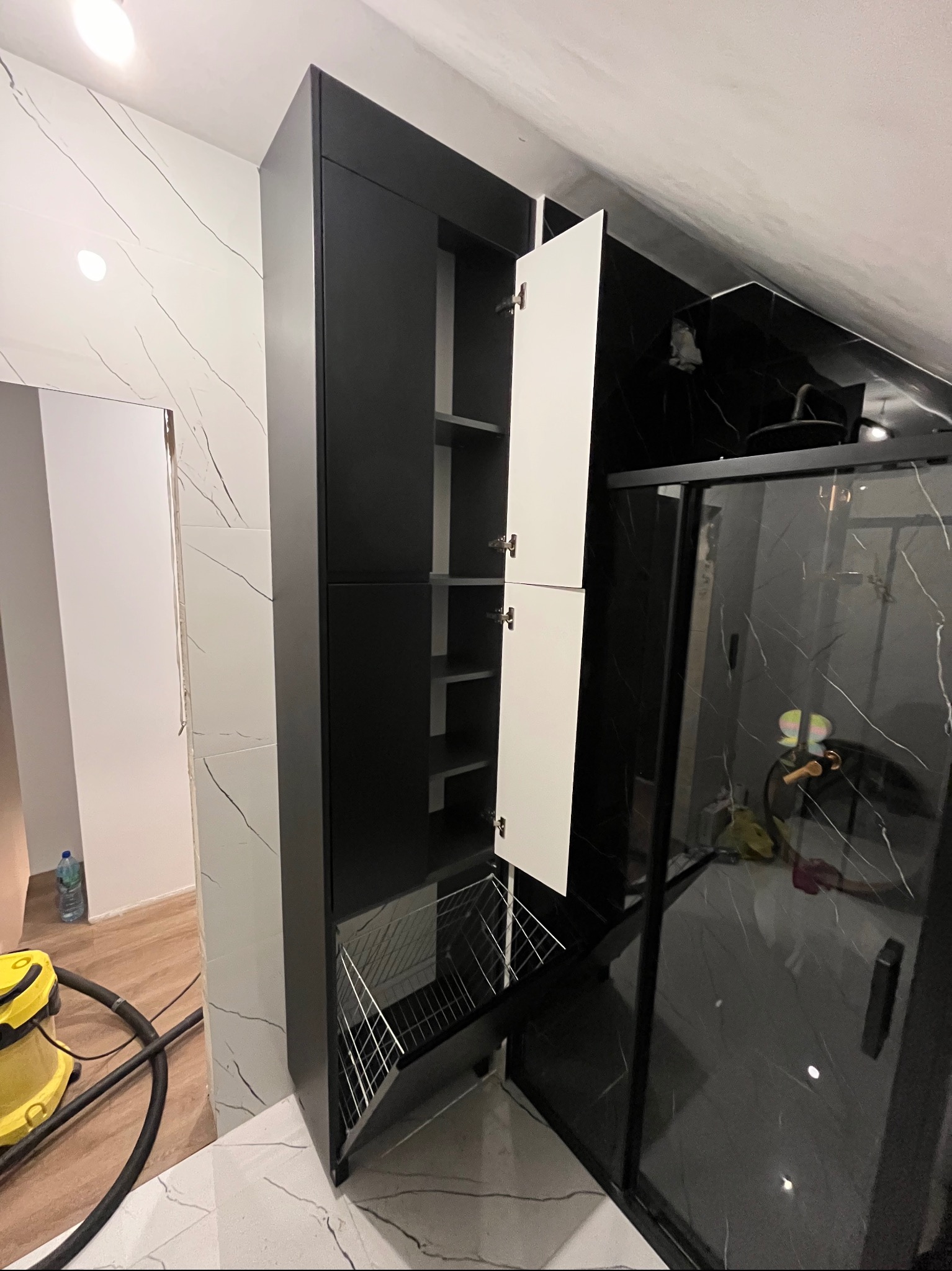 Wysoka, czarno-biała szafa wnękowa z otwartymi białymi drzwiczkami i metalowym koszem na dole, w łazience z czarno-białymi płytkami imitującymi marmur, widoczny fragment kabiny prysznicowej...