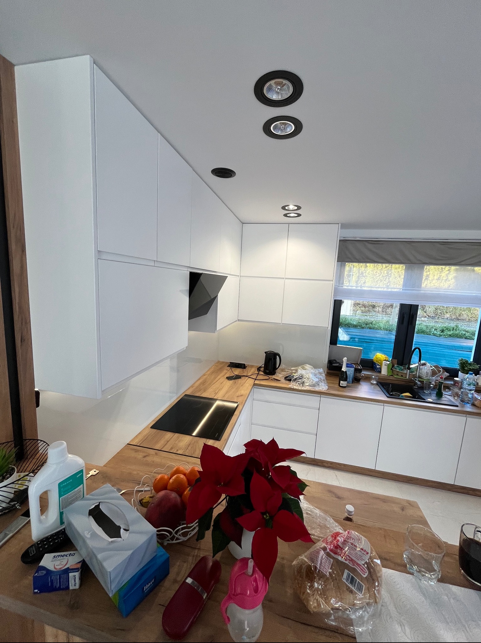 Nowoczesna kuchnia z białymi szafkami, drewnianym blatem i widocznymi elementami wyposażenia, takimi jak płyta indukcyjna i okap. Na pierwszym planie, na blacie, znajdują się artykuły spożywcze...