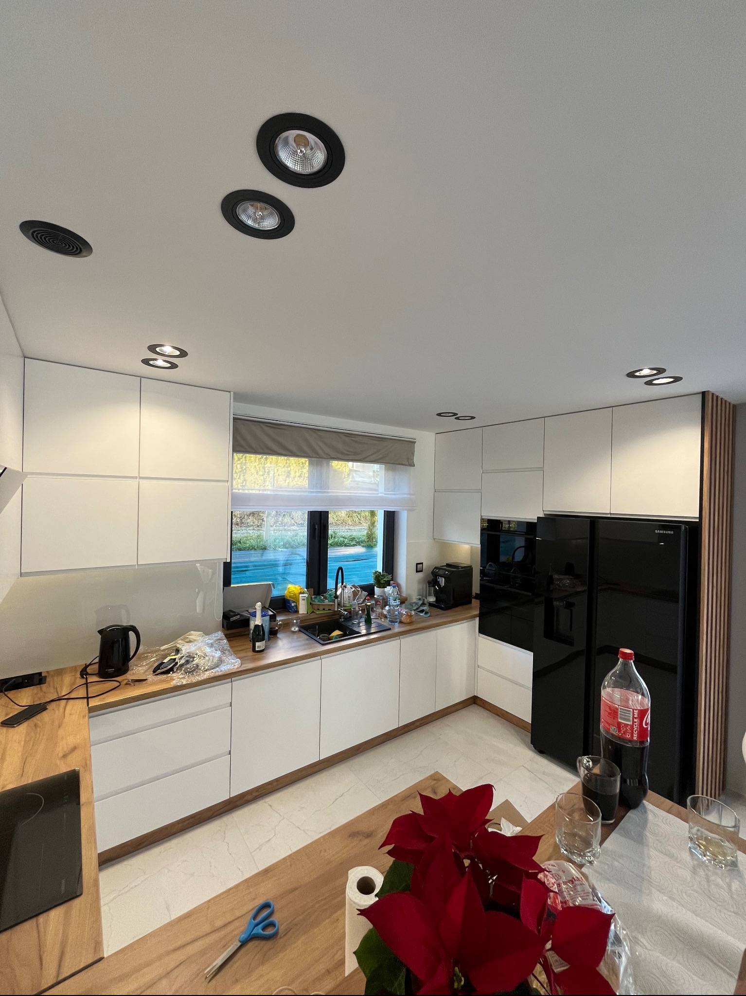 Nowoczesna kuchnia z białymi szafkami, drewnianym blatem i czarną lodówką Samsung, widok na okno z roletą, na stole czerwona poinsecja i butelka Coca-Coli.