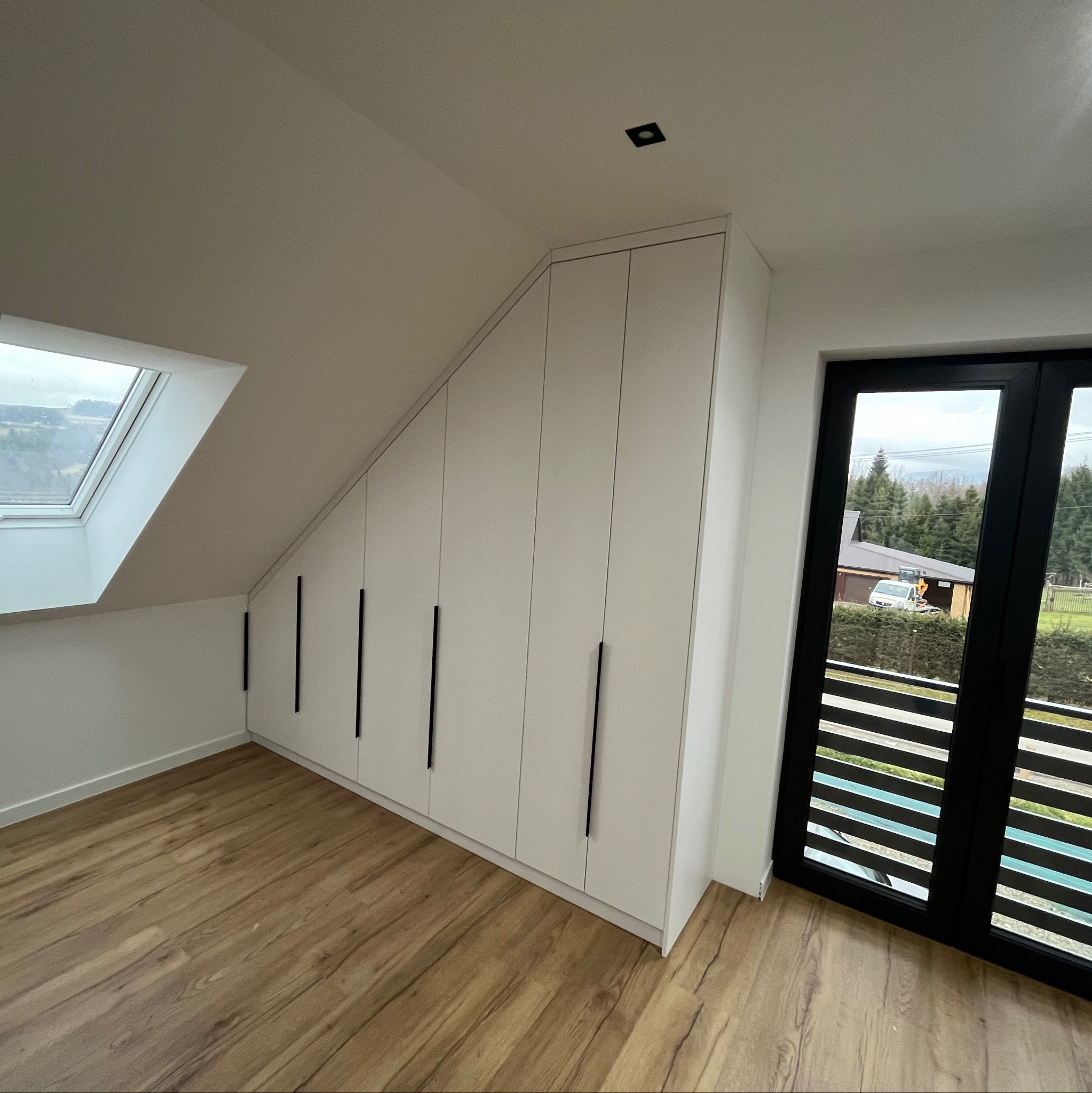 Biała szafa wnękowa pod skosem dachu z długimi czarnymi uchwytami, widok na okno dachowe i przesuwne okno balkonowe z widokiem na zewnątrz.