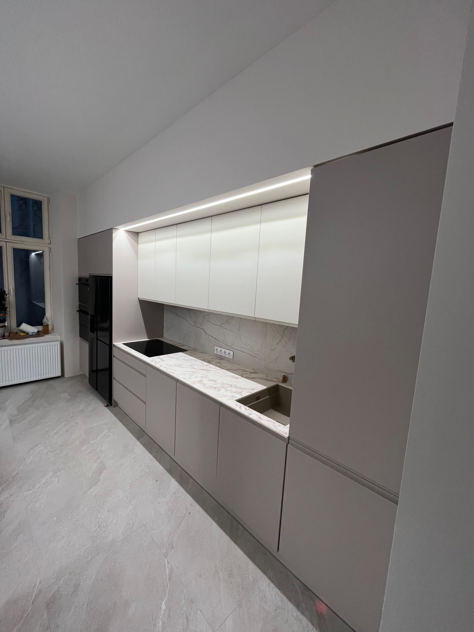 Nowoczesna kuchnia z zabudową w odcieniach szarości i bieli, z marmurowym blatem i oświetleniem LED, zrealizowana w Sułkowicach. Widoczne okno z grzejnikiem w tle.