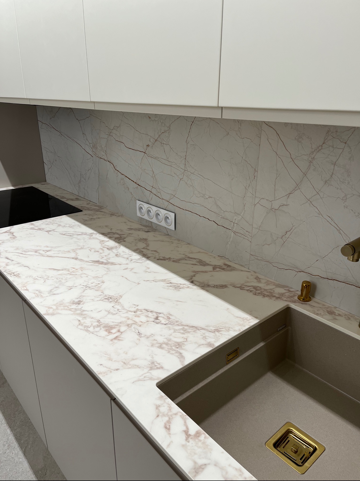 Fragment nowoczesnej kuchni z blatem imitującym marmur, szarym zlewozmywakiem z pozłacanym odpływem i białymi szafkami wiszącymi, uzupełniony o gniazdka elektryczne w białej ramce.