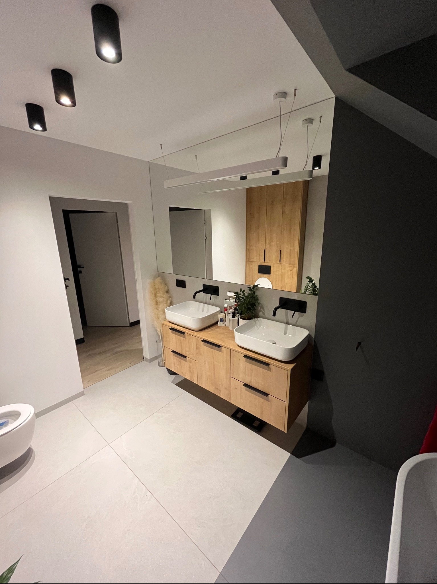 Nowoczesna łazienka z drewnianą szafką pod umywalki nablatowe, czarne baterie, minimalistyczne oświetlenie i lustro bez ramy.