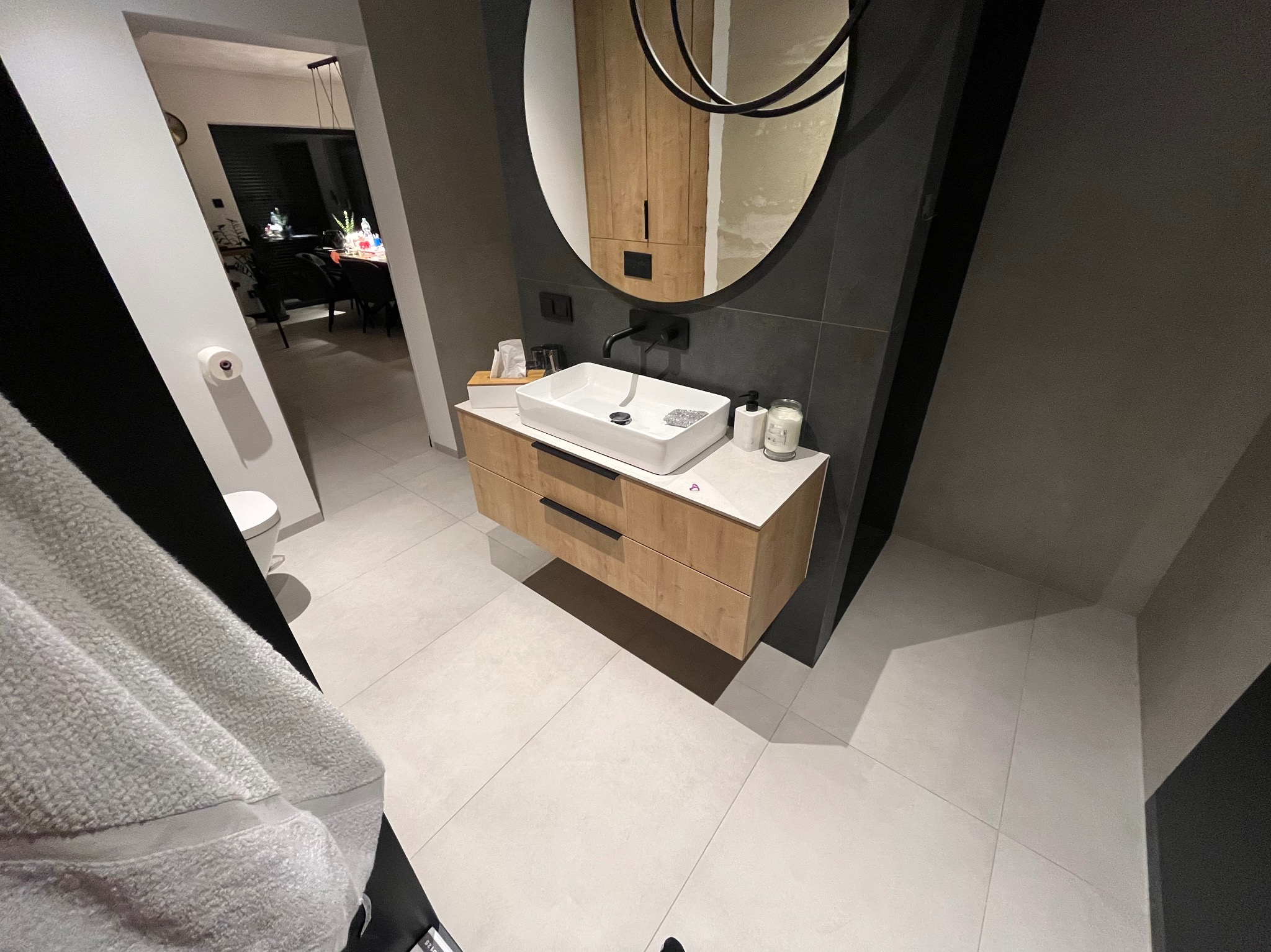 Nowoczesna łazienka z drewnianą szafką pod umywalką, białą umywalką nablatową, okrągłym lustrem z czarną ramą i czarną armaturą, widok z perspektywy żabiej