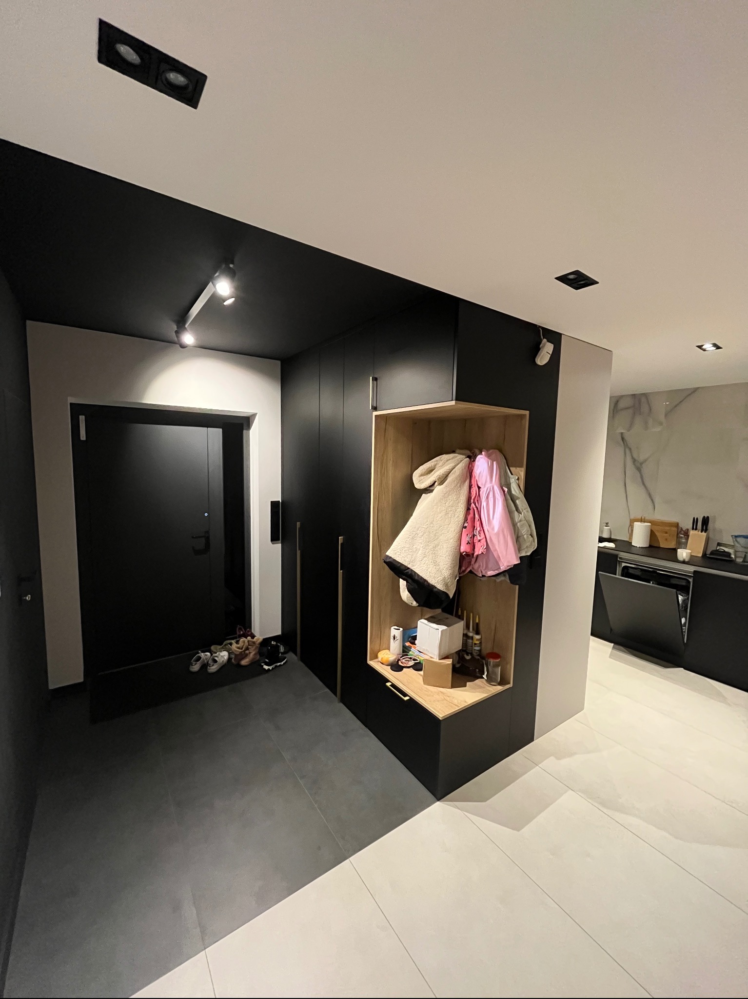 Nowoczesny przedpokój z czarną zabudową szafy wnękowej z otwartą półką na ubrania i akcesoria, widoczna część kuchni w tle.
