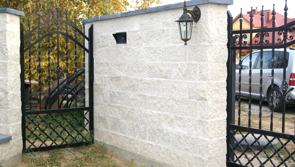 Murowane ogrodzenie z jasnych bloczków z czarną, kutą bramą i furtką, oświetlone zewnętrzną lampą, z widoczną skrzynką na listy.