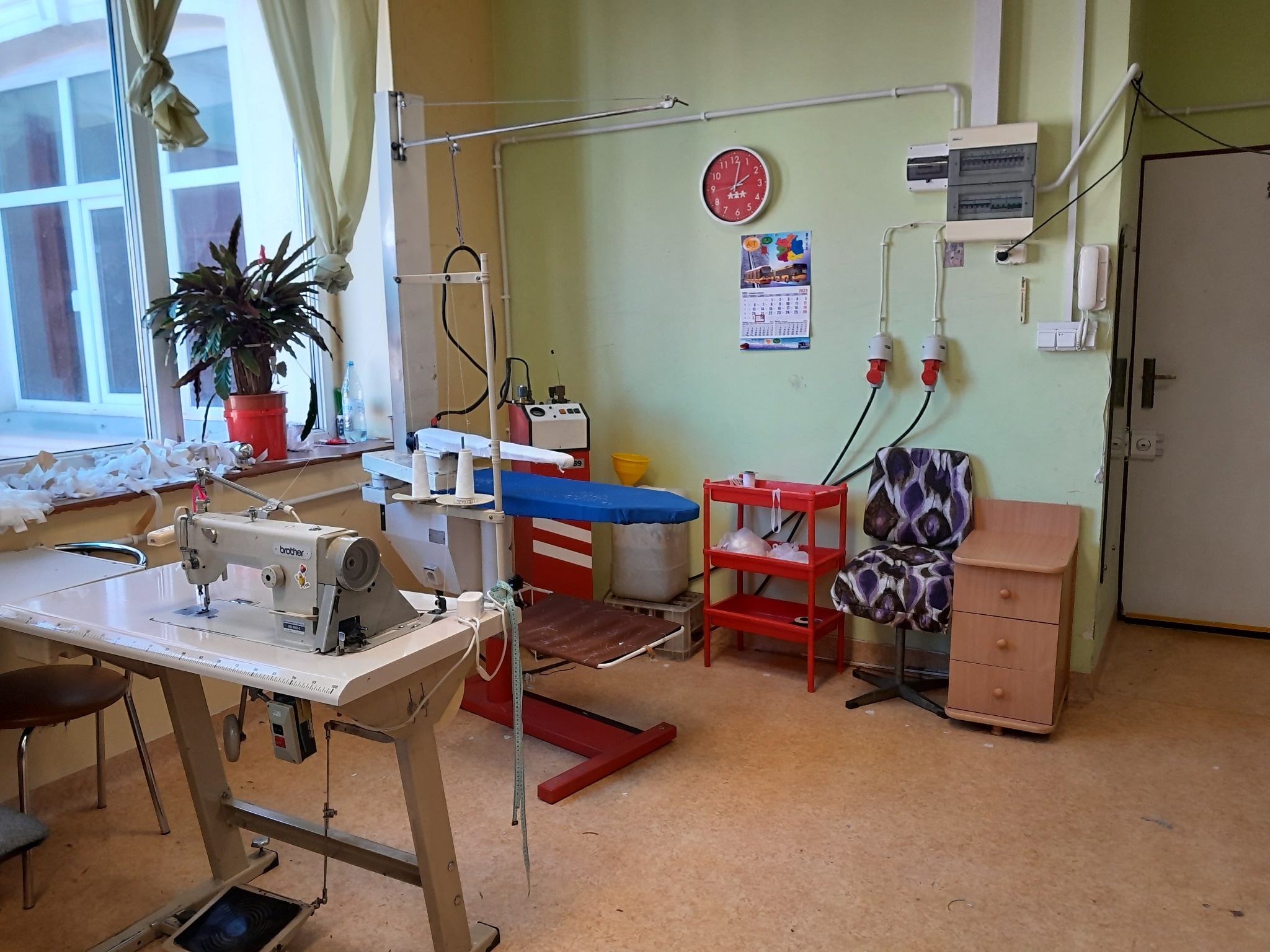 Wnętrze szwalni: maszyna do szycia Brother, deska do prasowania z niebieskim pokrowcem, czerwony regał i kalendarz na ścianie. Widok na okno z rośliną.