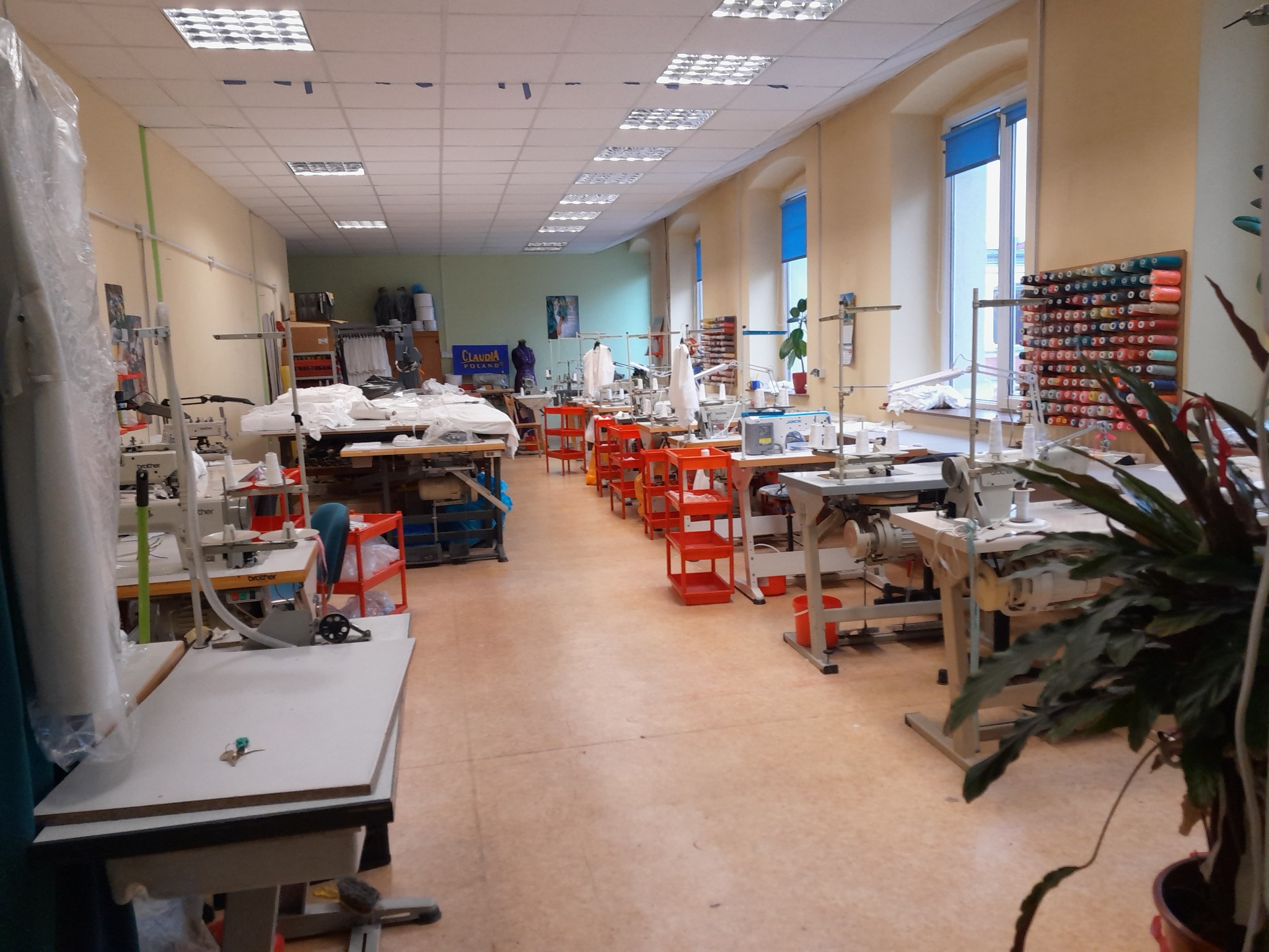 Jasne wnętrze szwalni z maszynami do szycia, stołami roboczymi i regałem z kolorowymi nićmi. Widok na przestronne pomieszczenie z oknami.