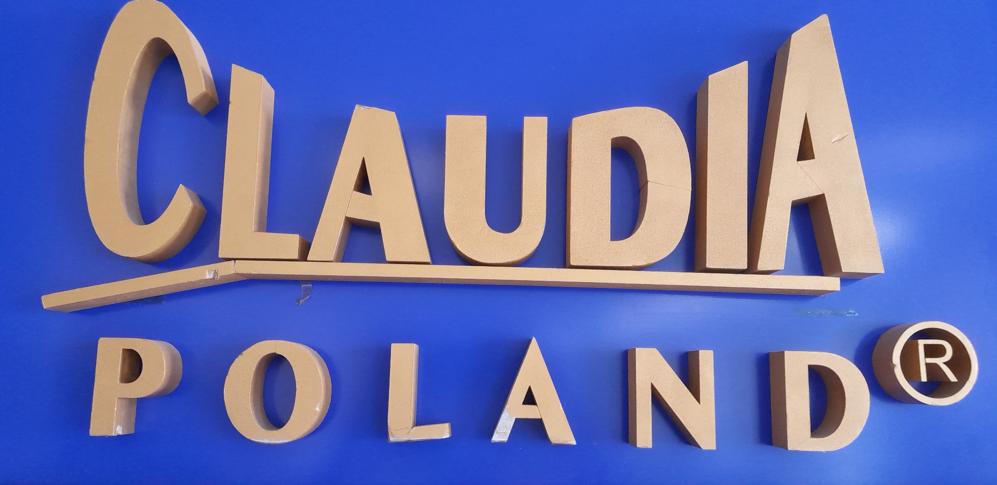 Złote, przestrzenne litery 'CLAUDIA POLAND®' na błękitnym tle. Widoczne niedoskonałości montażu i drobne uszkodzenia liter.
