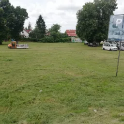 Świeżo skoszone pole z pomarańczowym wozem serwisowym w tle, znak parkingu 'Pod Zamkiem' po prawej, drzewa i budynki w oddali.