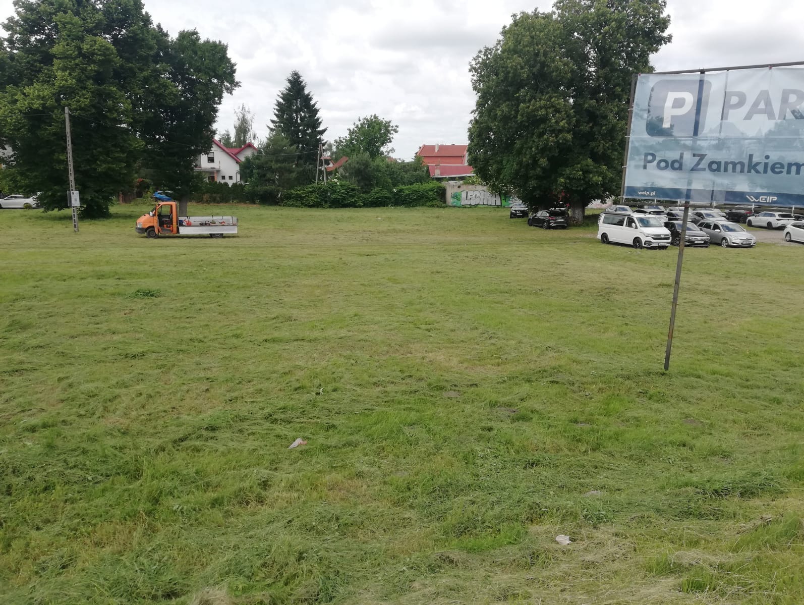 Świeżo skoszone pole z pomarańczowym wozem serwisowym w tle, znak parkingu 'Pod Zamkiem' po prawej, drzewa i budynki w oddali.