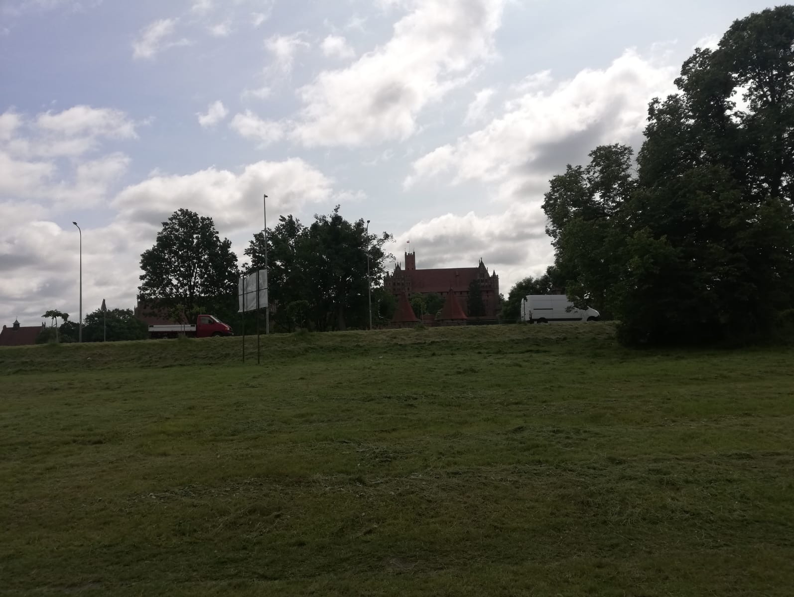 Świeżo skoszona trawa na tle zamku w Malborku, widoczny czerwony samochód i biały van. Pochmurne niebo z białymi obłokami.