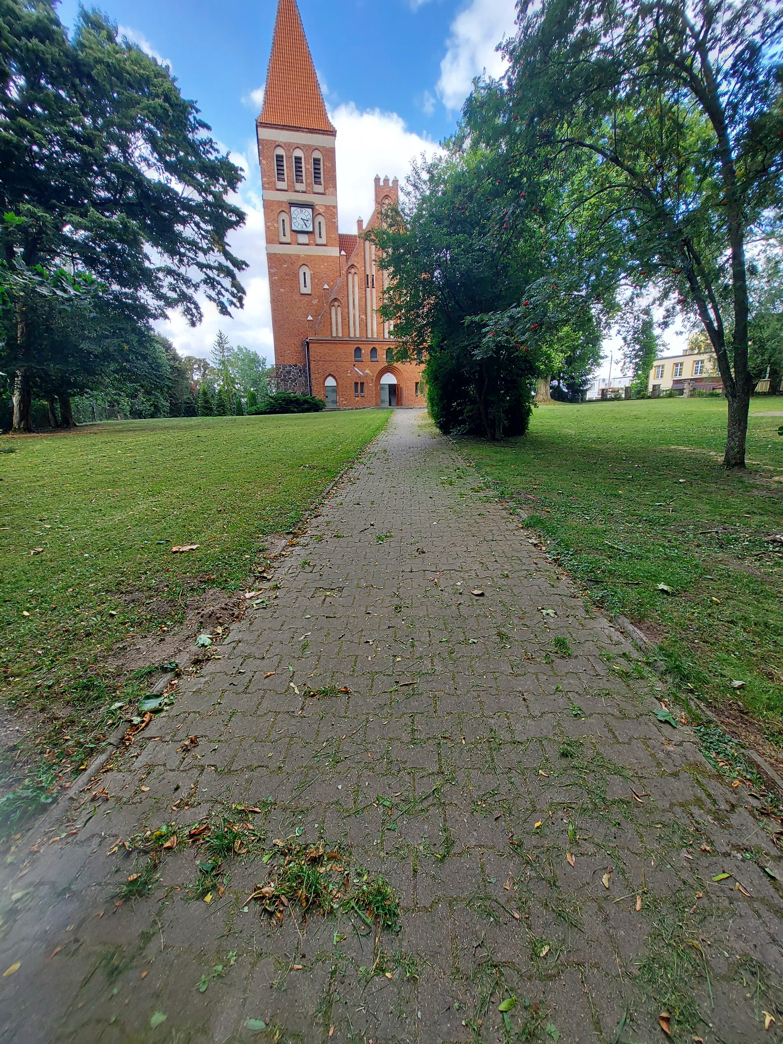 Aleja z kostki prowadzi do ceglanego budynku z wieżą zegarową, otoczonego zielenią i drzewami. Świeżo skoszona trawa leży na kostce. Błękitne niebo z chmurami w tle.