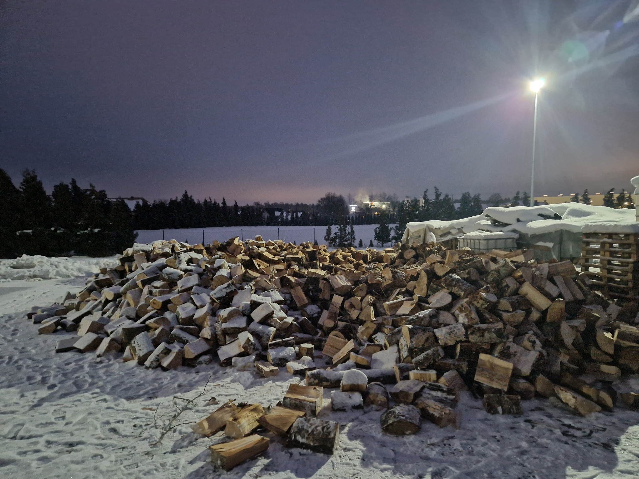 Stos drewna kominkowego pokryty śniegiem, oświetlony lampą uliczną na tle zimowego krajobrazu o zmierzchu. Drzewa w tle.
