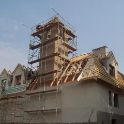 Budynek w trakcie budowy z widoczną konstrukcją więźby dachowej, rusztowaniami i wieżą wznoszącą się ponad dachem, częściowo pokrytym dachówką. Widoczne elementy izolacji na dachu.