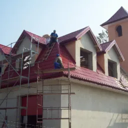 Budowa domu z wieżą: czerwona dachówka ułożona na części dachu, rusztowania wokół budynku, dwóch robotników na dachu, okna poddasza bez szyb.