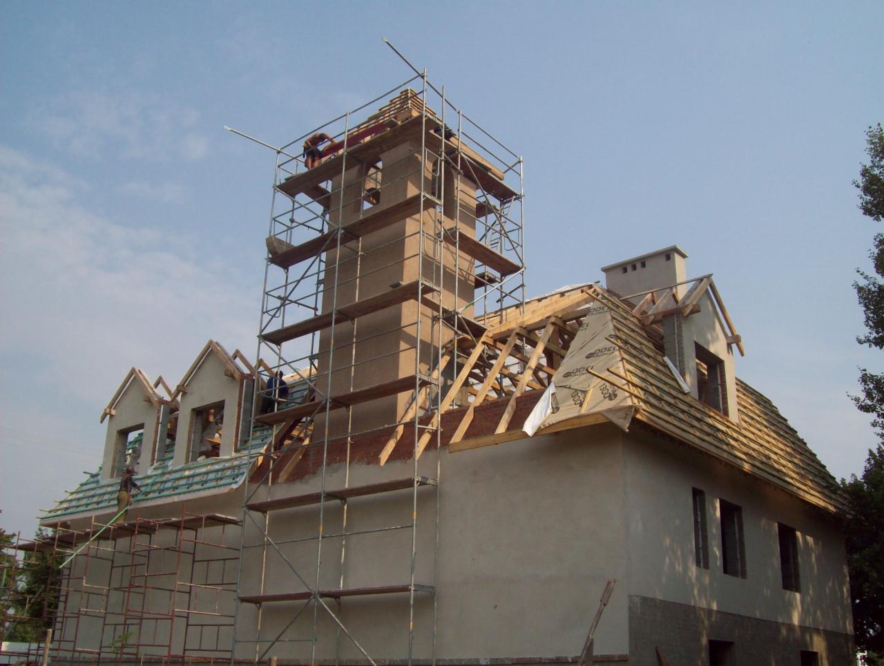 Budynek w trakcie budowy z widoczną konstrukcją więźby dachowej, rusztowaniami i wieżą wznoszącą się ponad dachem, częściowo pokrytym dachówką. Widoczne elementy izolacji na dachu.