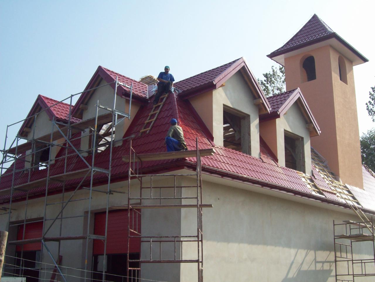 Budowa domu z wieżą: czerwona dachówka ułożona na części dachu, rusztowania wokół budynku, dwóch robotników na dachu, okna poddasza bez szyb.