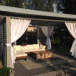 Drewniana altana z białymi zasłonami, rattanowym narożnikiem i stolikiem na kostce brukowej, otoczona zielenią. Widok na zewnątrz z elementami ogrodu.