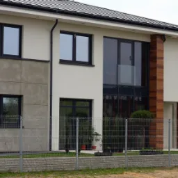 Nowoczesny dom jednorodzinny z dużymi oknami w czarnych ramach, z widocznym ogrodzeniem z siatki i betonową podmurówką.