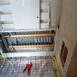 Rozdzielacz ogrzewania podłogowego z zaworami termostatycznymi, rurami i narzędziami (obcinak do rur, klucz nastawny) na folii izolacyjnej z nadrukowaną siatką, widok z góry podczas montażu.