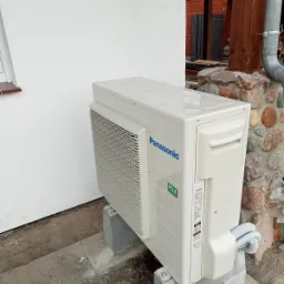 Zewnętrzna jednostka klimatyzacji Panasonic R32, zamontowana na betonowych bloczkach przy ścianie budynku z widocznym fragmentem drewnianej konstrukcji werandy.