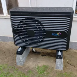 Czarna jednostka zewnętrzna z poziomymi paskami, marki FOX AIR, zamontowana na betonowych wspornikach obok ściany budynku.