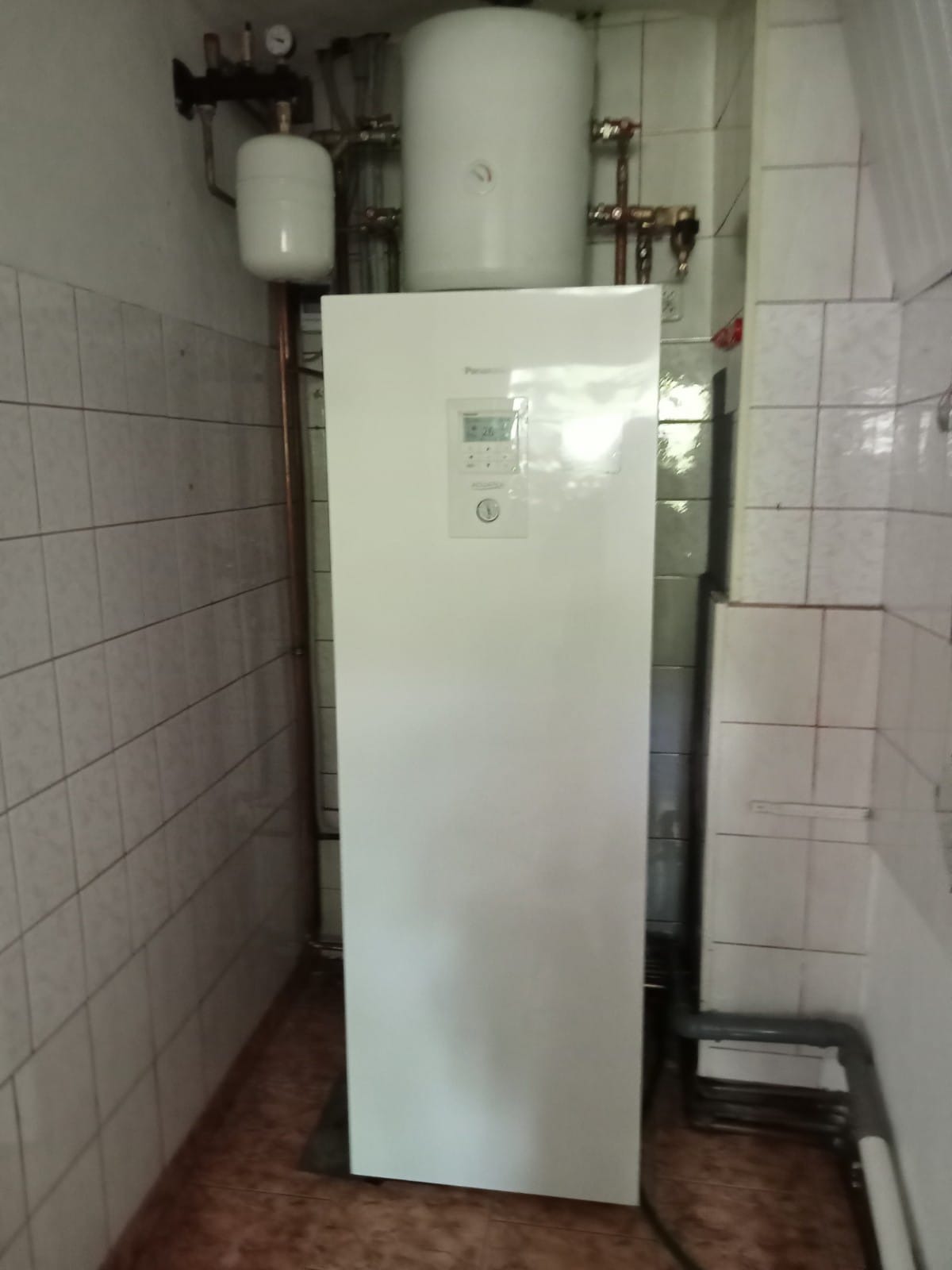 Biała pompa ciepła Panasonic z wyświetlaczem ustawień, zainstalowana w pomieszczeniu z wykafelkowanymi ścianami i widocznymi rurami instalacji.