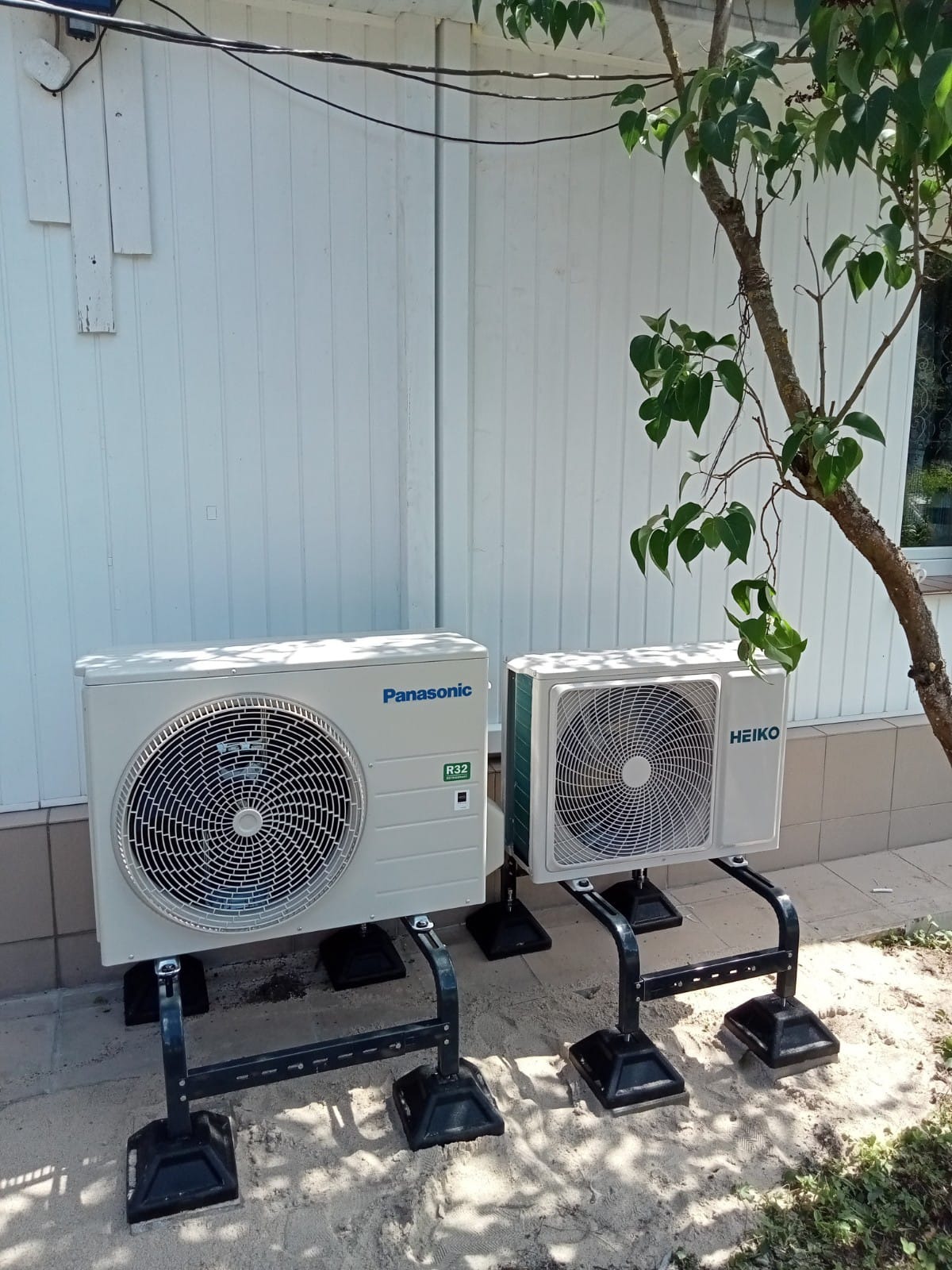 Dwa zewnętrzne moduły klimatyzacji różnych marek (Panasonic i Heiko) zamontowane na metalowych wspornikach przed budynkiem z białym sidingiem, z widocznym drzewem z liśćmi po prawej stronie.
