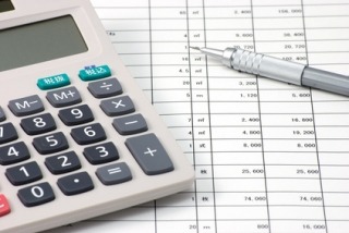 Kalkulator i długopis leżące na arkuszu kalkulacyjnym z danymi finansowymi w Łącku. Widoczny fragment tabeli z liczbami i nagłówkami kolumn.