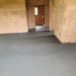 Wnętrze budynku w stanie surowym z wylaną, gładką, szarą posadzką jastrychową, ściany z czerwonej cegły dziurawki, widoczne przejścia do kolejnych pomieszczeń.