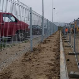 LECHDOM GARWOLIN - Długa linia stalowego ogrodzenia panelowego na nowo przygotowanym terenie, z widocznymi samochodami i słupami oświetleniowymi w tle, oraz pracownikiem w pomarańczowej kamizelce w oddali.