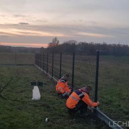LECHDOM GARWOLIN - Dwóch monterów w pomarańczowych kurtkach mocuje metalowe panele do betonowej podmurówki na tle zachodzącego słońca i rozległego pola.