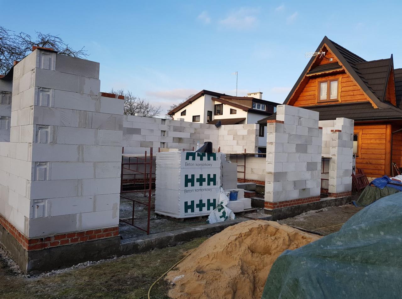 Wznoszenie ścian domu z betonu komórkowego H+H na fundamencie z ceglaną podmurówką, widoczny stos zapakowanych bloczków, rusztowanie, piasek i dom w tle.