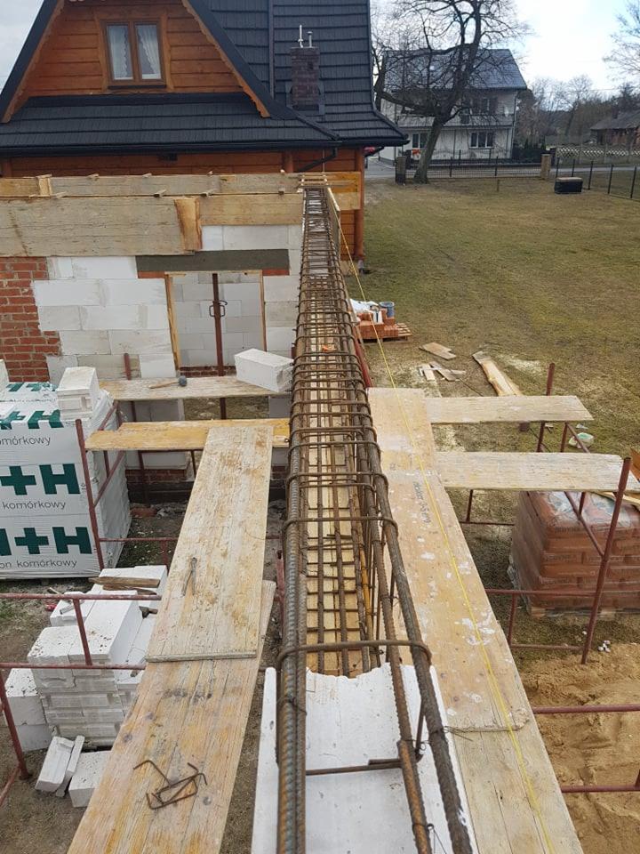 Zbrojenie pod wieniec na budowie domu z białego pustaka komórkowego, widok z góry na drewniane stemple i deskowanie, w tle dom z brązowego drewna i trawnik.