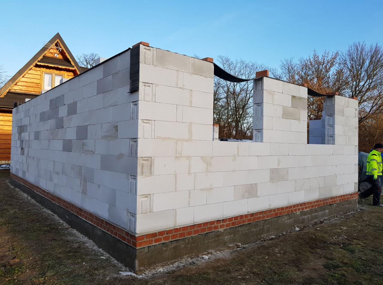 Wznoszenie ścian parteru domu z betonu komórkowego na fundamencie z cegły, widoczny fragment drewnianego domu w tle.