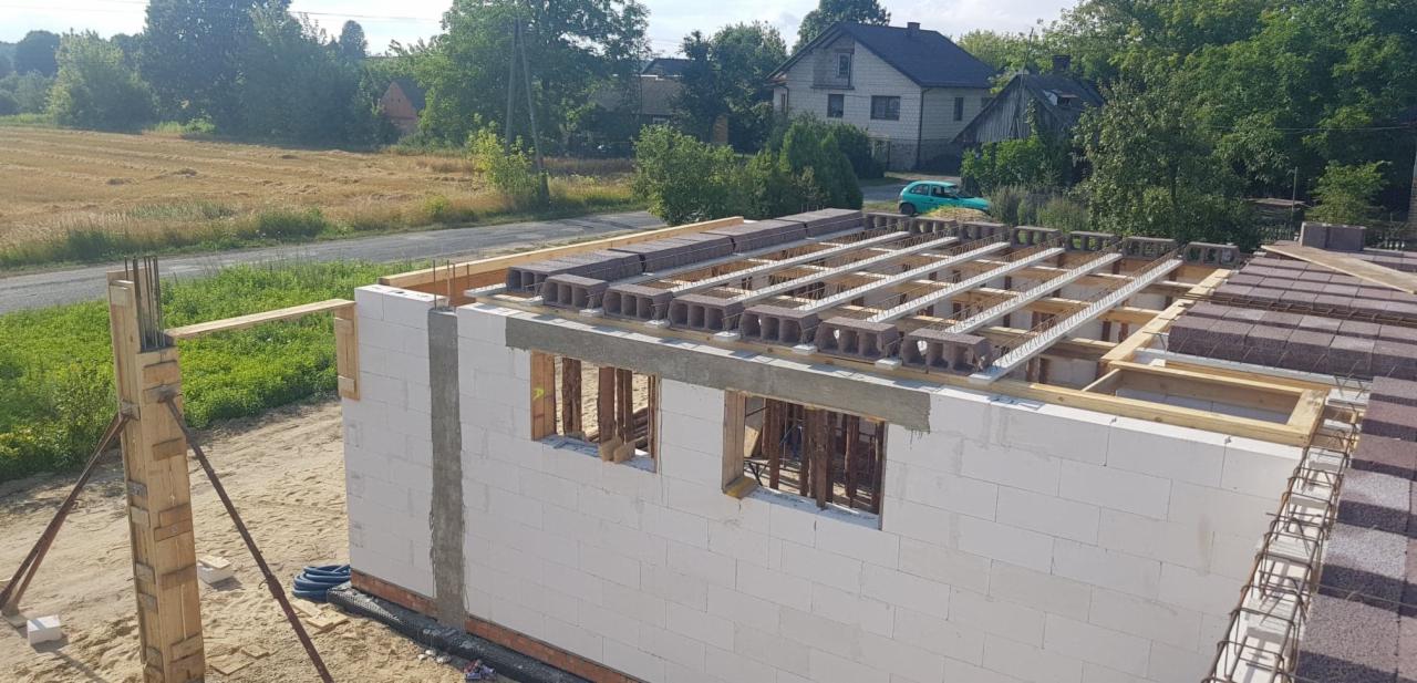 Budowa domu jednorodzinnego: wznoszenie ścian z bloczków, widoczne zbrojenia i elementy stropu gęstożebrowego, w tle zielone drzewa i zabudowa wiejska.