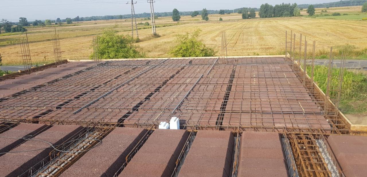 Zbrojenie stropu Teriva na budowie domu jednorodzinnego, widok z góry na ułożone bloczki i siatkę z prętów stalowych, wystające pionowe pręty, w tle pole i drzewa.