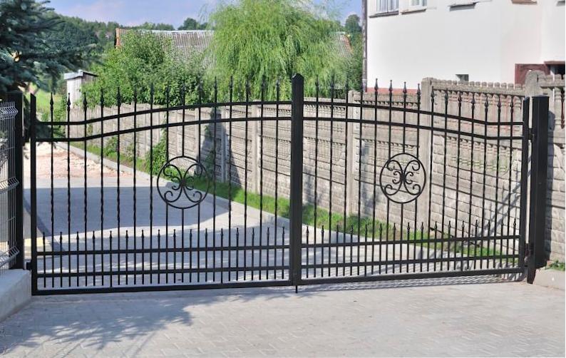 Czarna, metalowa brama wjazdowa z ozdobnymi elementami na tle betonowego ogrodzenia i zieleni.