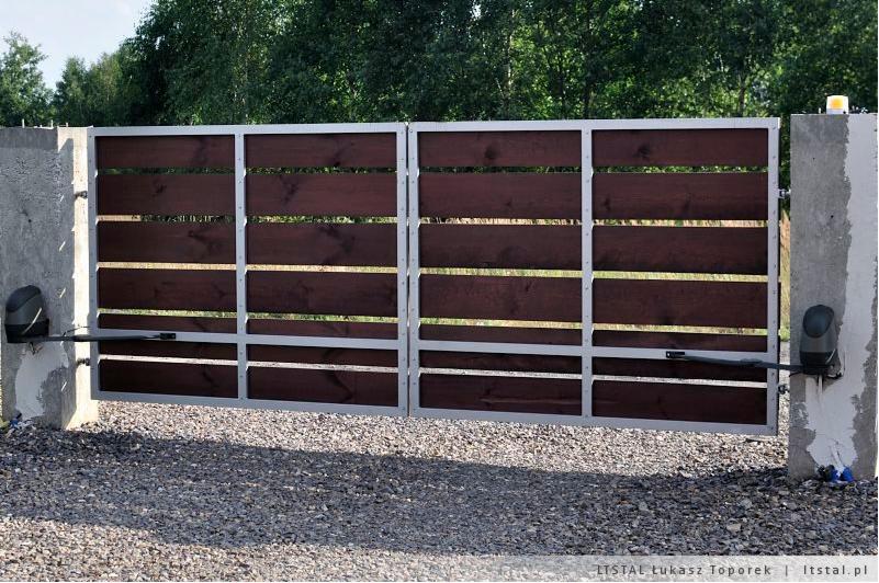 Dwuskrzydłowa brama wjazdowa z poziomymi deskami z ciemnego drewna, osadzona w metalowej ramie, z automatyką do otwierania, zamontowana na betonowych słupkach na tle zieleni.
