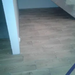 Ułożona podłoga z płytek imitujących drewno, widoczne białe krzyżyki dystansowe, fragment schodów w tle.