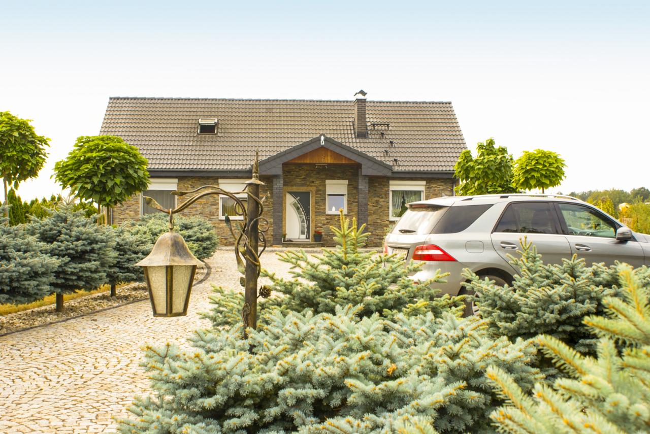 Posesja z domem jednorodzinnym, podjazdem z kostki brukowej, ozdobnymi krzewami iglastymi, lampą ogrodową z kutego metalu i zaparkowanym samochodem osobowym.