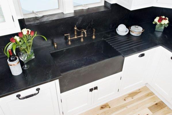 Czarny kamienny zlew farmerski z mosiężną armaturą na tle białych szafek kuchennych, uzupełniony o kwiaty, wino i drewnianą podłogę.