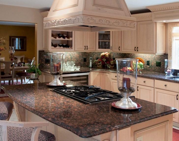 Jasna kuchnia z brązowym blatem granitowym, wyspą kuchenną z płytą grzewczą, dekoracyjnym okapem i szklanymi lampionami ze świecami.