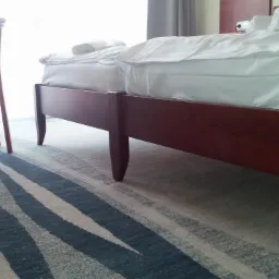 Wzorzysta, szaro-biała podłoga w pokoju hotelowym z drewnianym łóżkiem i krzesłami. Wykładzina dywanowa z abstrakcyjnym motywem fal. Jasne, naturalne światło.